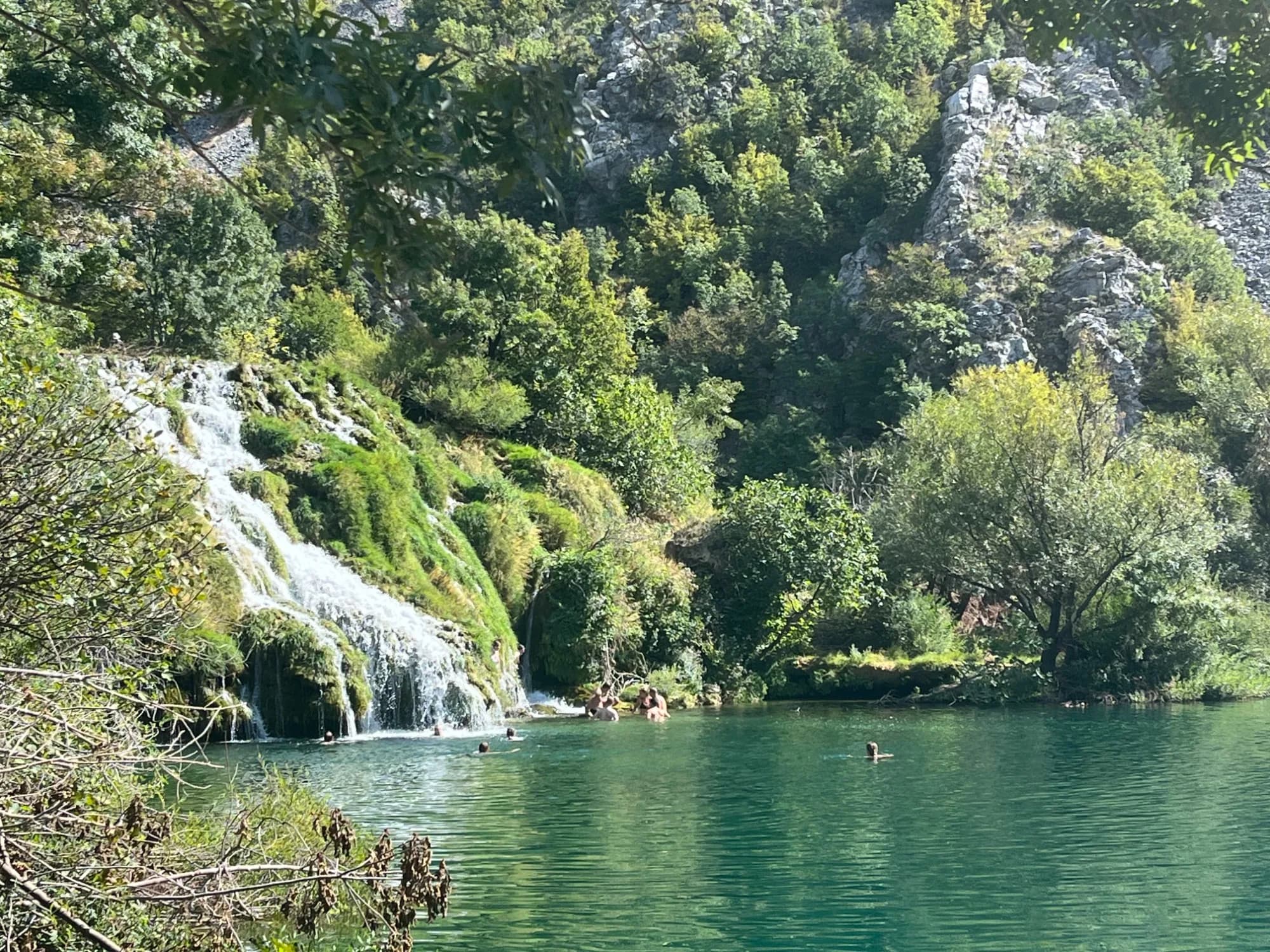Wasserfall stürzt über bemooste Felsstufen in einen türkisfarbenen Fluss, in dem Menschen baden